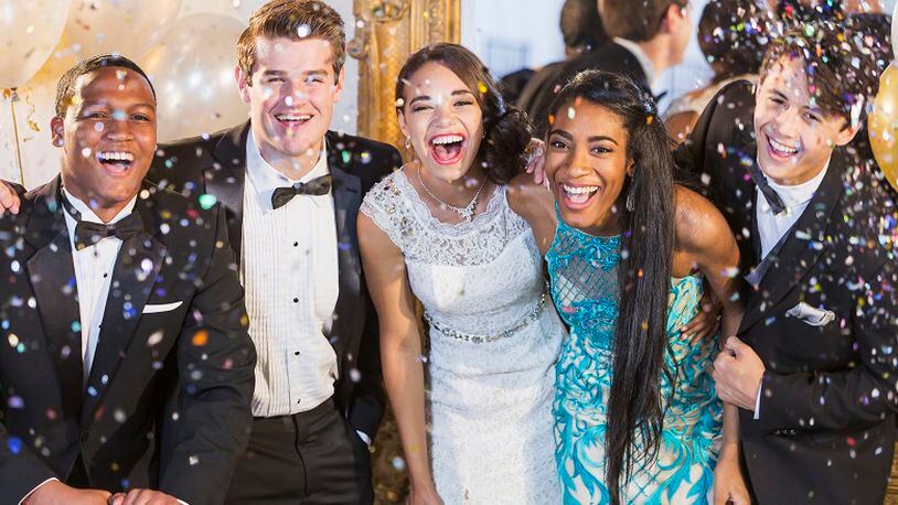 A group of five multi-ethnic teenagers and young adults dressed in formalwear - dresses and tuxedos. They are at a special event, a party or prom, standing together in a row, laughing and smiling. Or maybe a New Year's Eve party, confetti in the air to celebrate.