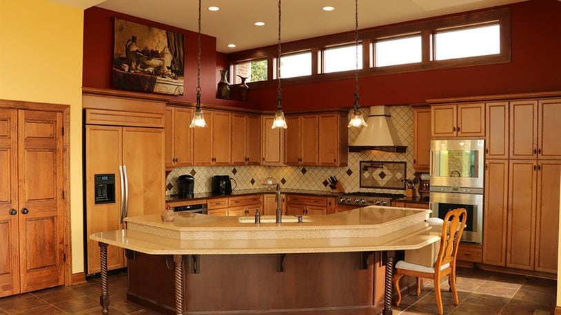 A wide, semi-circular island with a surrounding tiered seating area is placed at the entrance to the kitchen. Under a row of clerestory windows, plant shelf, recessed ceiling and hanging lights, nut-stained oversized cabinets wrap around the work space with quartz countertops and tiled backsplashes.