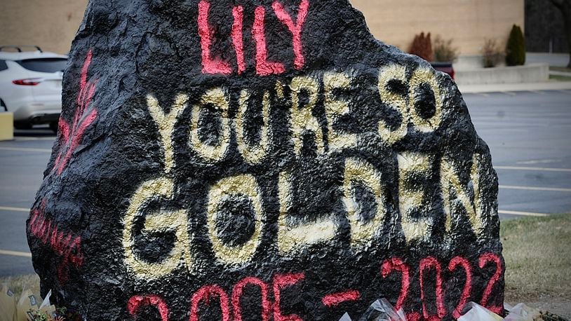 A large rock painted with "Lily, you're so golden, 2005-2022" in red and yellow spraypaint, with a memorial of flowers surrounding the base.