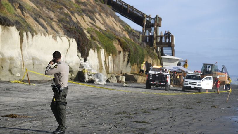 A San Diego County Sheriff's deputy looks on as search and rescue personnel work at the site of a cliff collapse at a popular beach Friday, Aug. 2, 2019, in Encinitas, Calif. The collapse killed at least three people and injured two.
