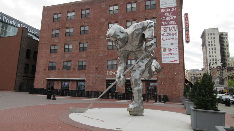 This May 11, 2017 shows "The Iron Man" sculpture of a hockey player next to the Prudential Center where the New Jersey Devils hockey team plays in Newark. In the background, signs can be seen for Dinosaur Bar-B-Cue BBQ and for Rock Plaza Lofts luxury apartments. Riots scarred Newark 50 years ago this summer, but tourism officials are hoping to attract more visitors as the city charts its comeback. (AP Photo/Beth J. Harpaz)