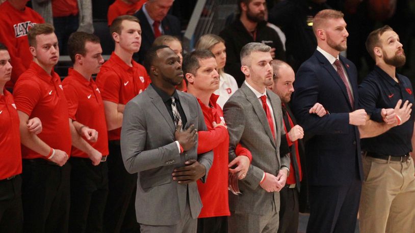 Dayton coaches, trainers and managers stand for the national anthem before a game against Indiana State on Nov. 9, 2019, at UD Arena. In the front row, from left to right, are Khyle Marshall, Mike Mulcahey, Brett Comer, James Haring, Sean Damaska and Casey Cathrall. David Jablonski/Staff