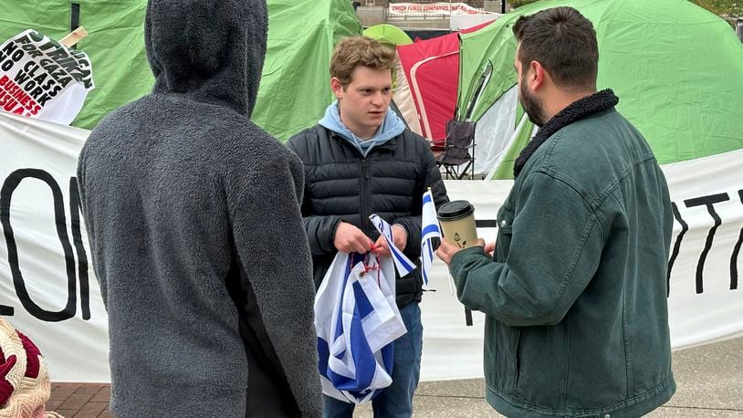 University of Michigan computer science junior Josh Brown, center, hands out miniature blue and white flags of Israel while standing Wednesday, April 23, 2024, in front of a banner reading “LONG LIVE THE INTIFADA," in Ann Arbor, Mich. The banner is part of a protest by students and groups demanding the Ann Arbor school divest from companies that do business with Israel. (AP Photo/Corey Williams)