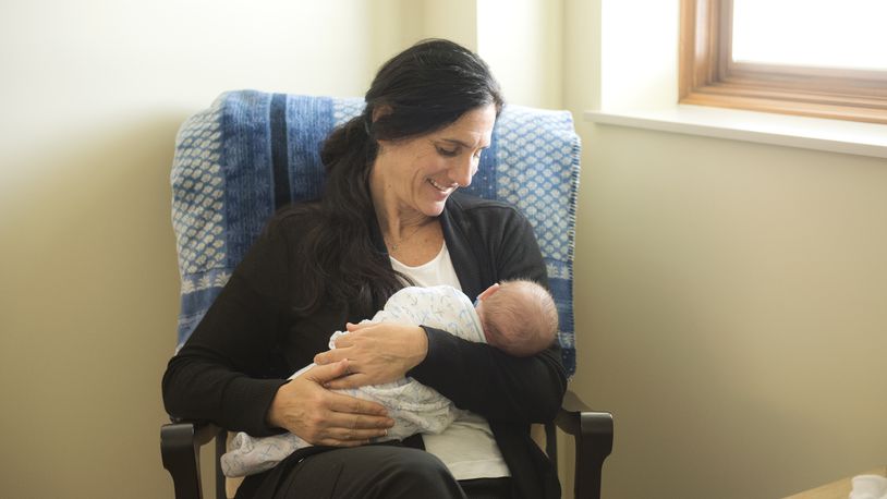 A smiling woman seated in a rocking chair cradles a newborn infant.