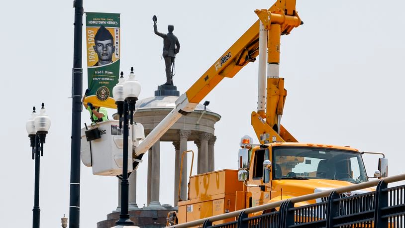 City crews install Hometown Heroes banners along the High Main Bridge with the Butler County Soldiers, Sailors and Pioneers Monument in the background Wednesday, May 17, 2023 in Hamilton. NICK GRAHAM/STAFF