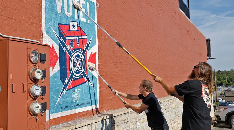 World famous artist Shepard Fairey, left, and his assistant paste up one of his "VOTE!" pictures on the wall at 128 East Main Street in Springfield Friday, Sept. 20, 2024. Fairey was in Ohio for ArtsVote Oho painting a mural in Cincinnati and before he headed back to California wanted to give a little positive light to Springfield with everything going on in the city. BILL LACKEY/STAFF