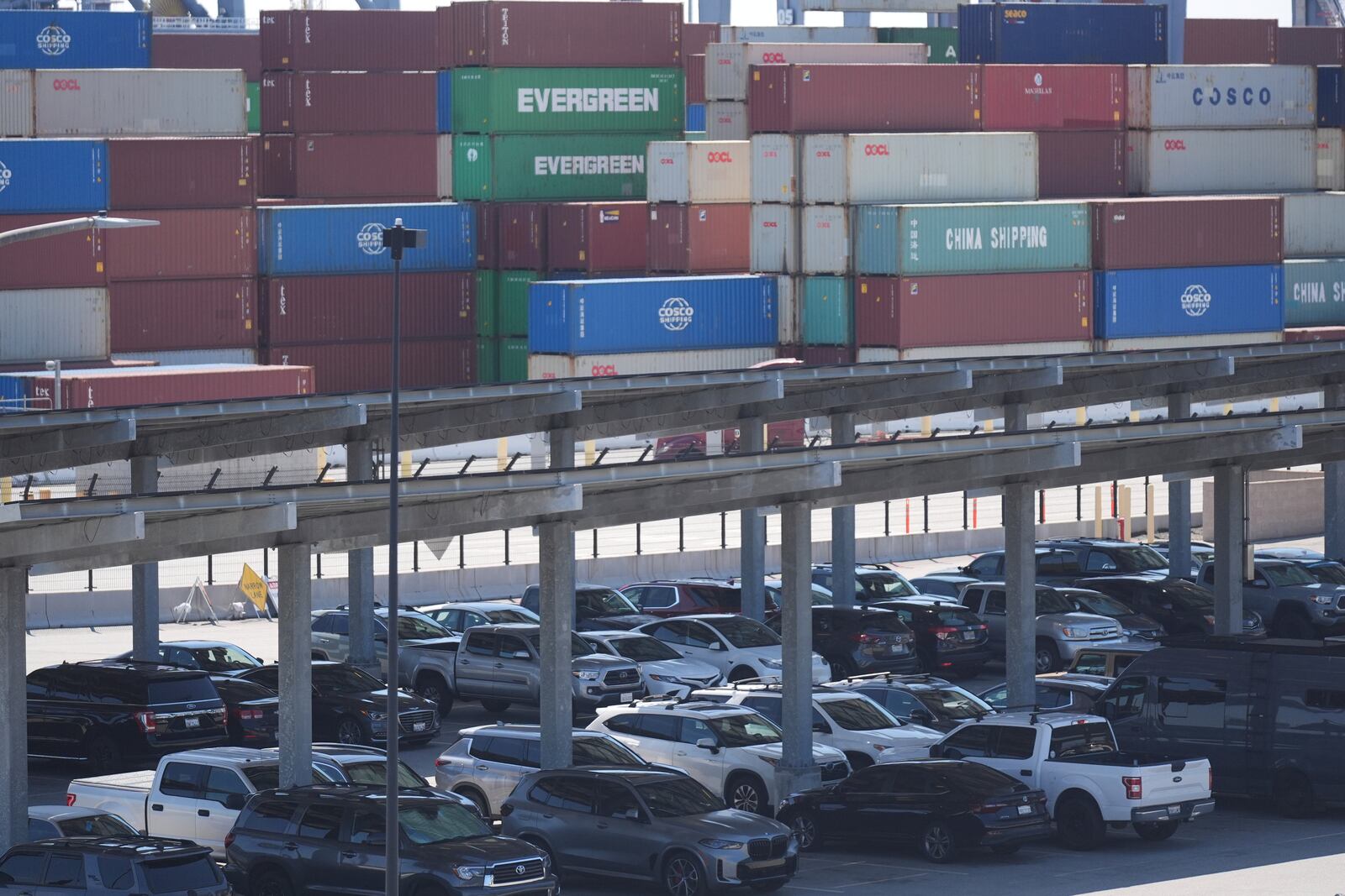 Vehicles are parked under solar panels as shipping containers sit stacked at the Long Beach Container Terminal in Long Beach, Calif., on Tuesday, April 8, 2025. (AP Photo/Damian Dovarganes)
