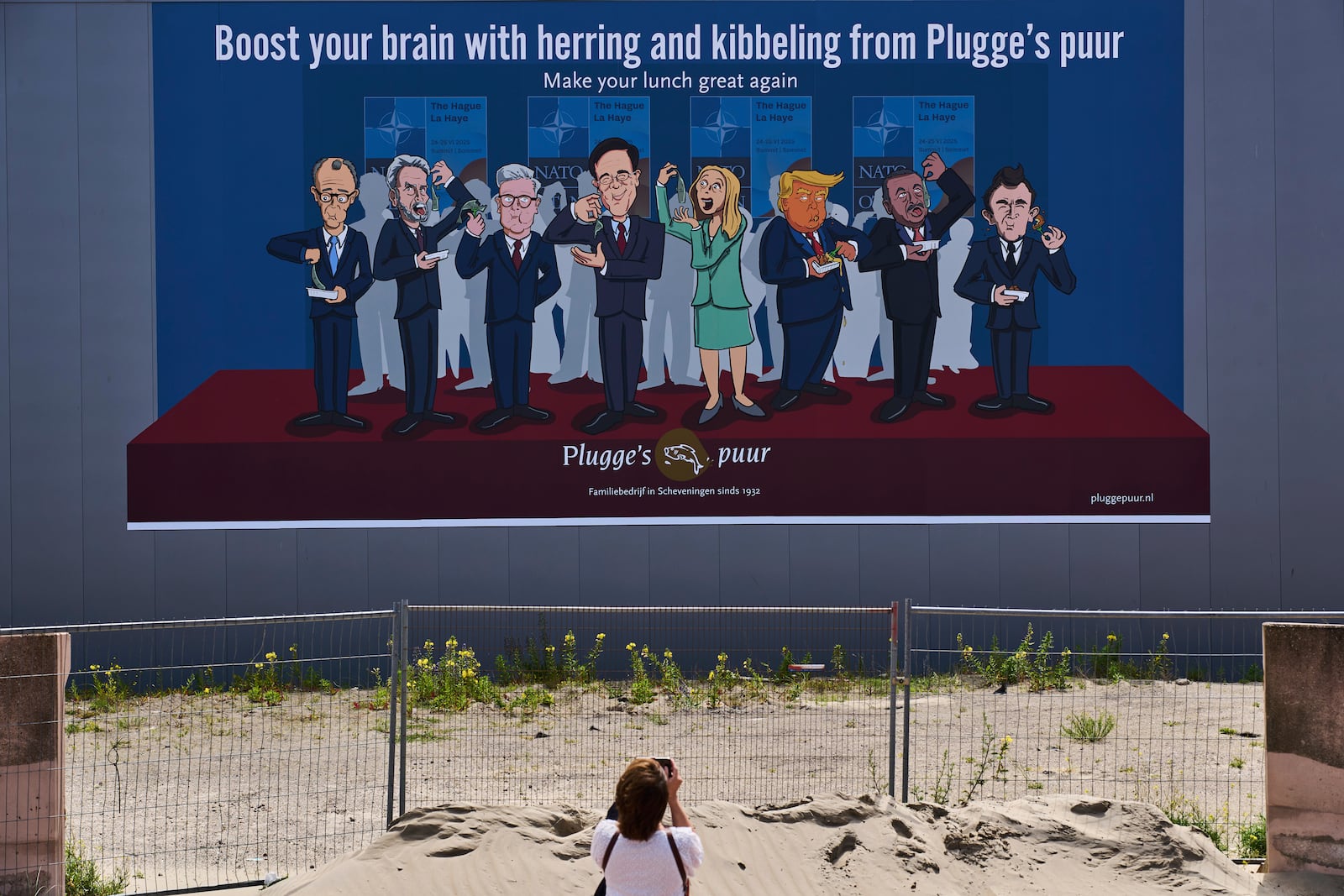 A woman takes a photo of a drawing showing World leaders from left, German Chancellor Friedrich Merz, Dutch Prime Minister Dick Schoof, British Prime Minister Keir Starmer, NATO Secretary General Mark Rutte, Italy's Prime Minister Giorgia Meloni, U.S. President Donald Trump, Turkish President Recep Tayyip Erdogan and French President Emmanuel Macron, eating herring and fried fish in The Hague, Netherlands, Sunday, June 15, 2025. (AP Photo/Peter Dejong)