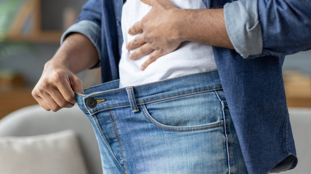 A man shows how much weight he has lost by holding out the waist of his jeans, symbolizing his successful diet. ISTOCK