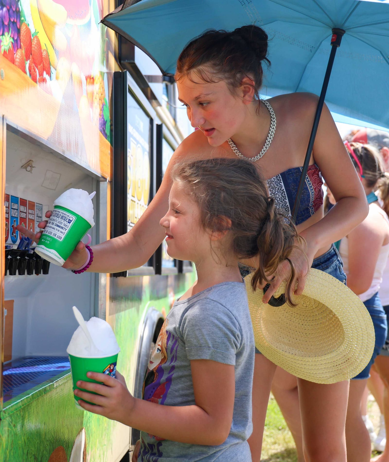 Taylor Baker (top), of Oxford, helps her sister Carmen Daniels, of Richmond, Ind., dispense flavoring in her cup of ice at a Kona Ice truck at the Centerpoint Energy Dayton Air Show on Sunday, June 22. The sisters were with their grandfather William Perry, of Richmond. William Perry's daughter Kyler, who is the aunt of the sisters, served in the U.S. Air Force. Kyler Perry died in 2022. BRYANT BILLING / STAFF