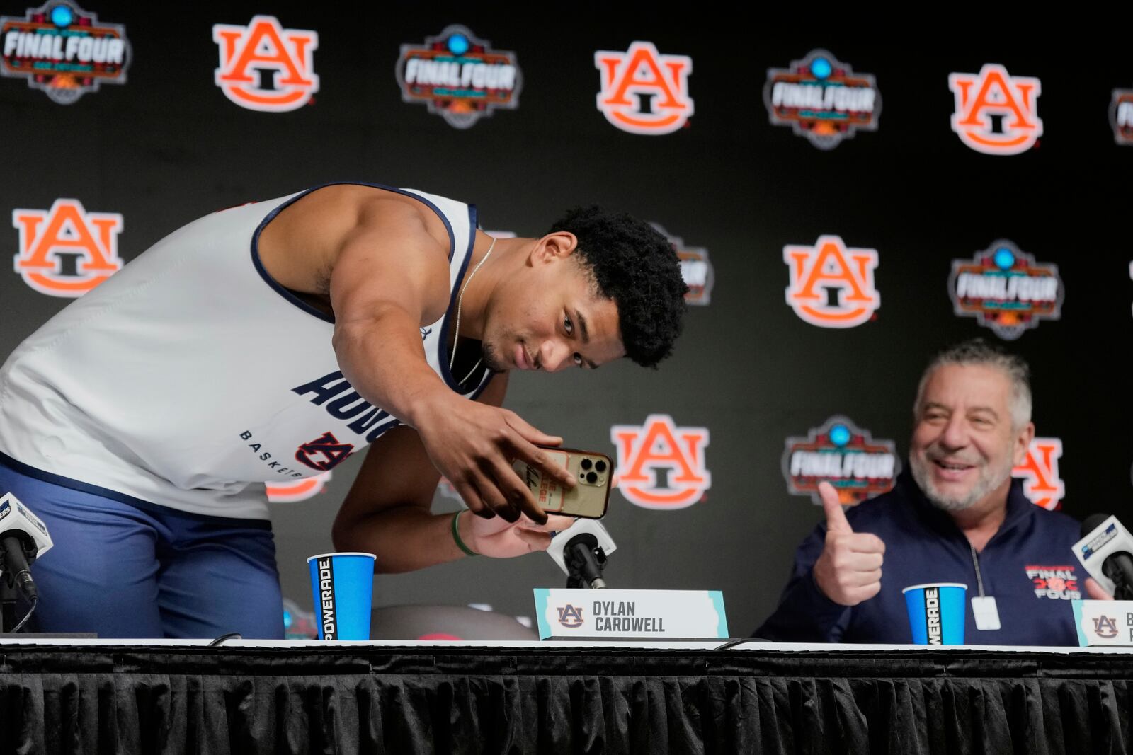 Auburn's Dylan Cardwell, left, takes a photograph of his nameplate as he joins head coach Bruce Pearl for a news conference at the Final Four of the NCAA college basketball tournament, Friday, April 4, 2025, in San Antonio. (AP Photo/Eric Gay)