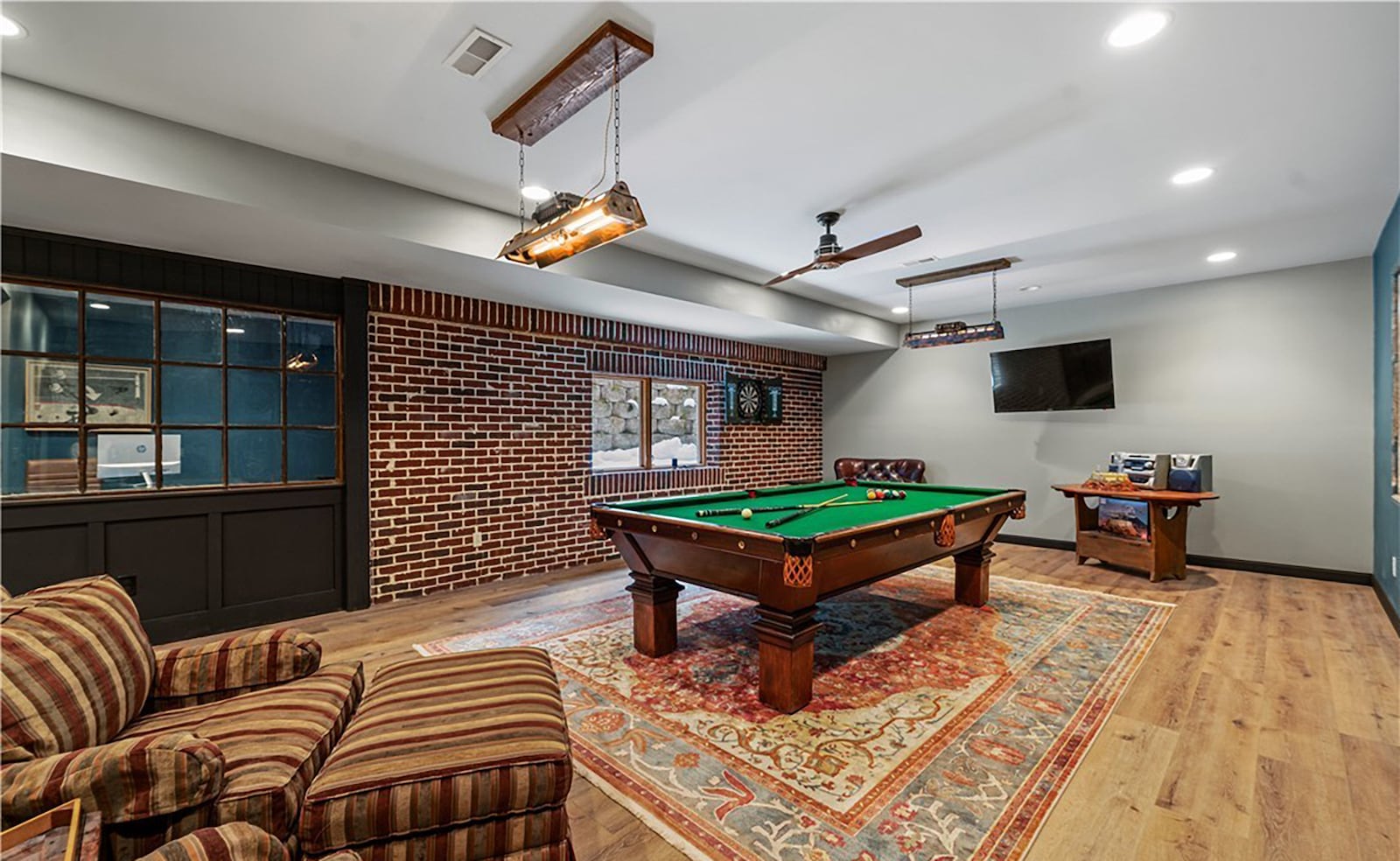 the finished basement features a recreation room with luxury vinyl plank flooring, a ceiling fan and two decorative fixtures and a brick wall accent. A window looks into a smaller attached billiard room. CONTRIBUTED