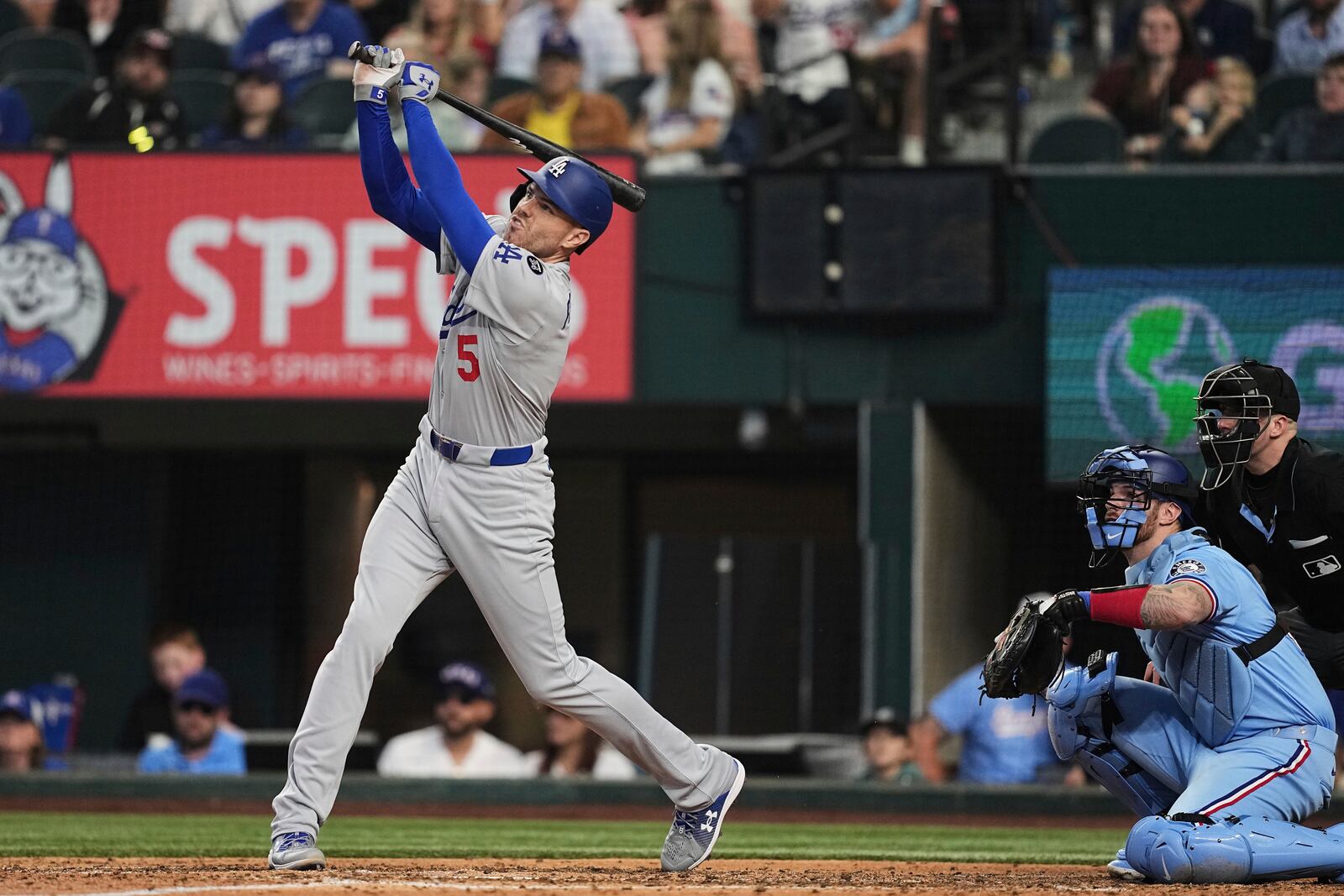 Los Angeles Dodgers' Freddie Freeman (5) follows through on a sacrifice fly that allowed Will Smith to score as Texas Rangers catcher Jonah Heim and umpire Nic Lentz, right, look on in the eighth inning of a baseball game in Arlington, Texas, Sunday, April 20, 2025. (AP Photo/Tony Gutierrez)