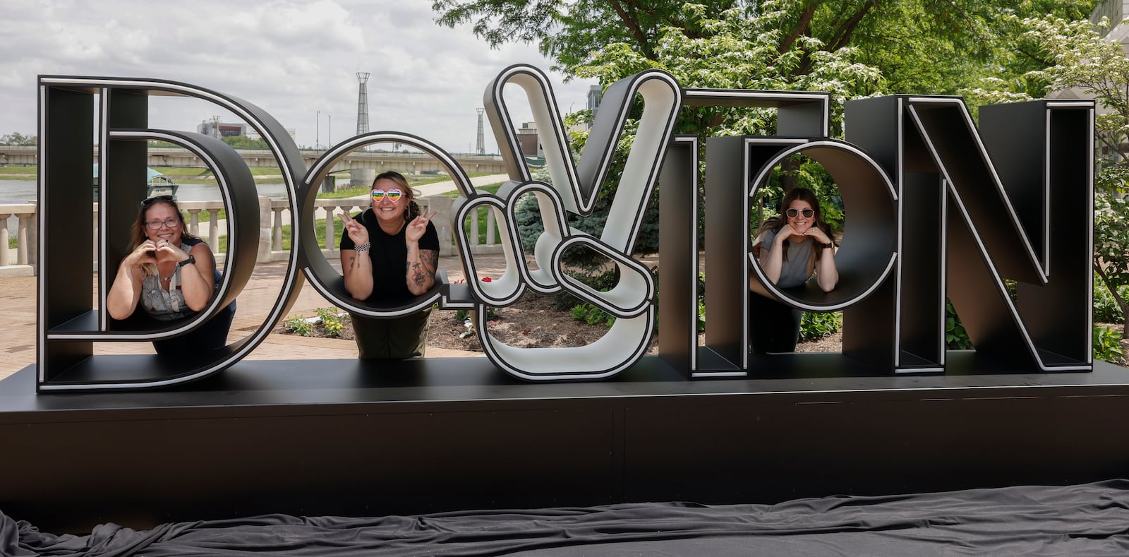 Left to right, SarahLydia Keihl (City of Dayton, Public Affairs), Jes Sands (Downtown Dayton Partnership), and Megan Peters (Cross Street Partners) take a photo at the newly unveiled Dayton Peace Sign on Thursday, May 15, 2025, at RiverScape MetroPark Festival Plaza. JOSEPH COOKE/STAFF