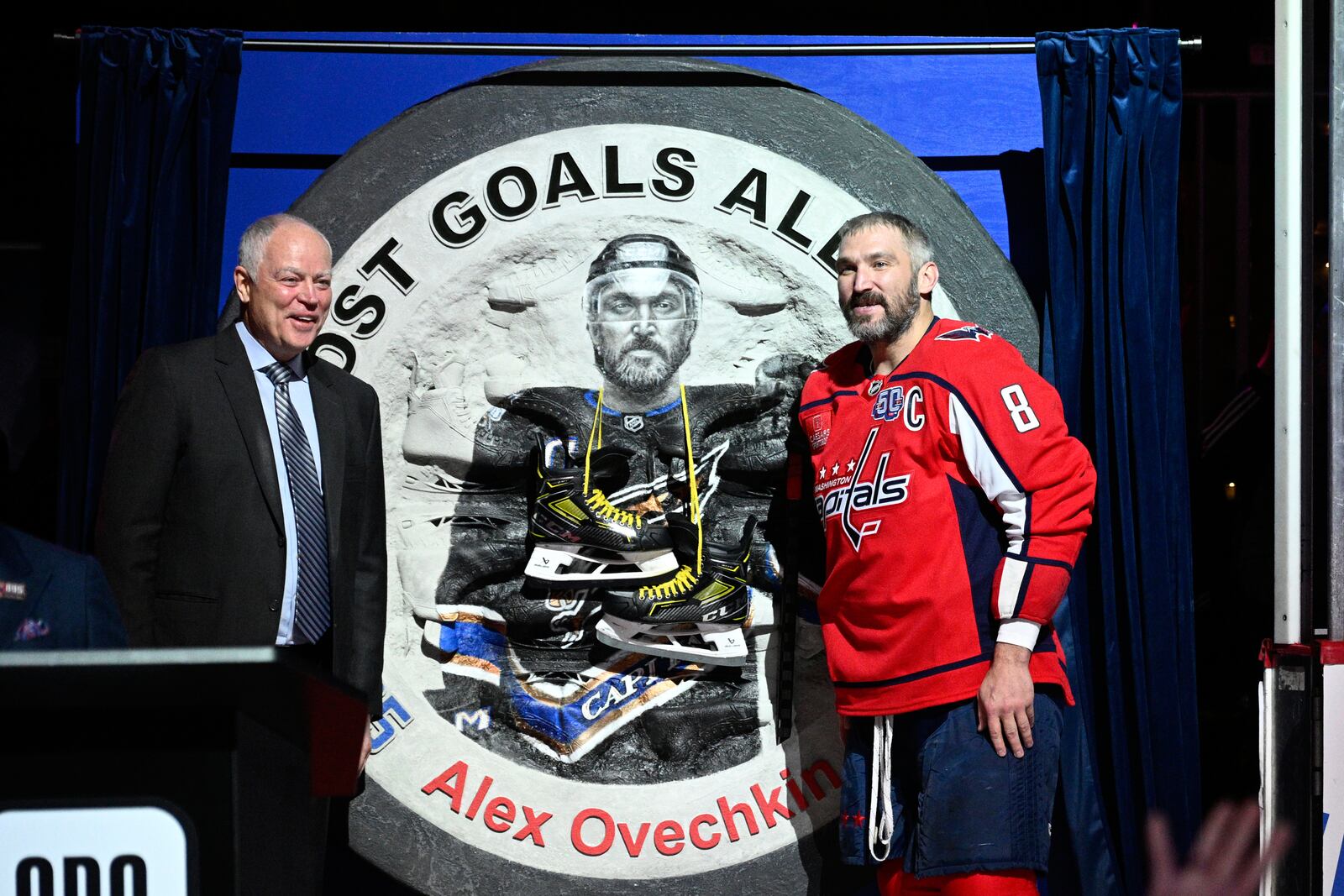 Washington Capitals left wing Alex Ovechkin (8) stands next to one of his gifts and Brian MacLellan, president of Capitals hockey operations, as he takes part in a ceremony to honor him for scoring 895 career NHL goals before an NHL hockey game against the Carolina Hurricanes, Thursday, April 10, 2025, in Washington. (AP Photo/Nick Wass)