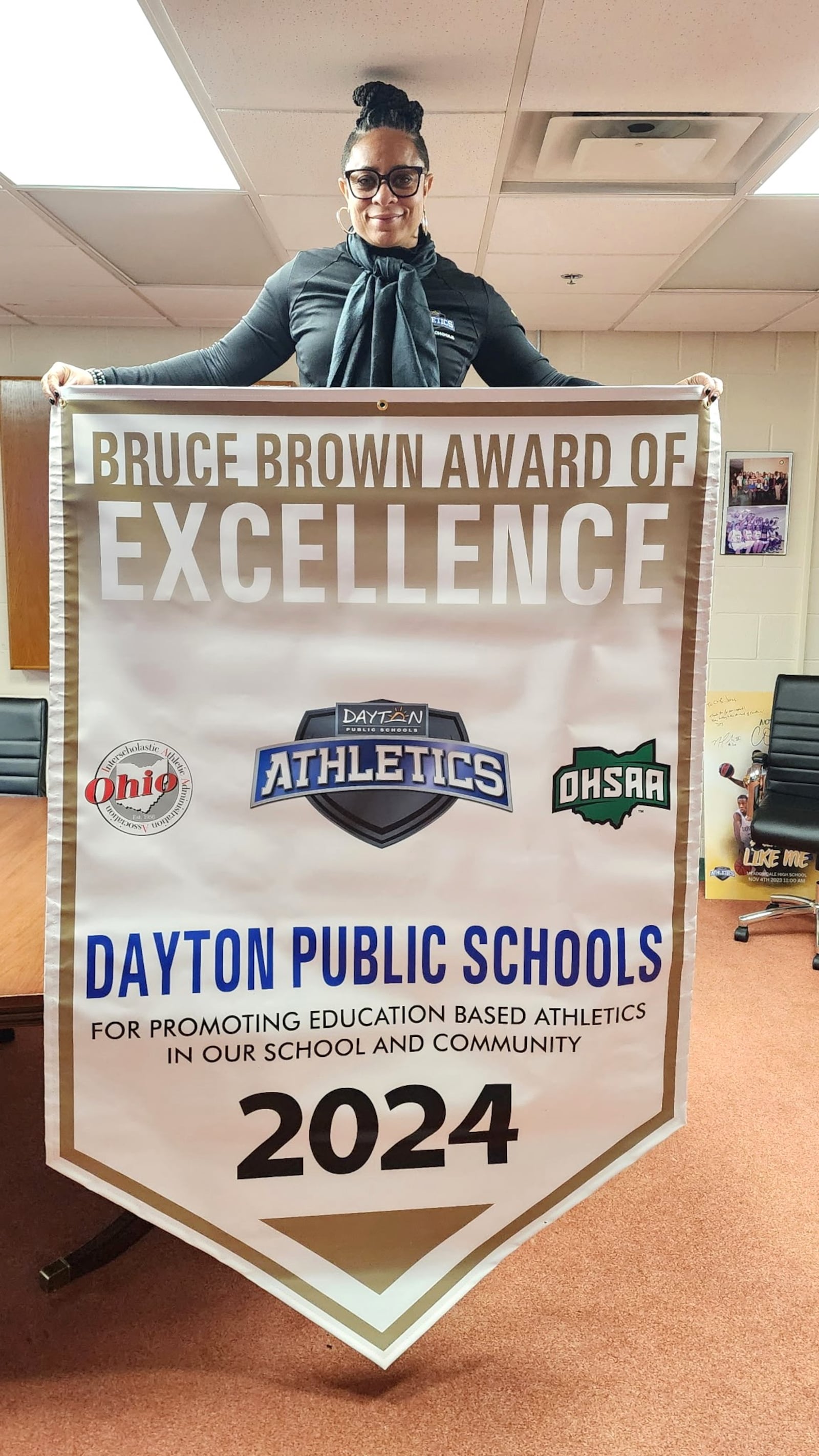 Victoria Jones,  the Director of Athletics for the Dayton Public Schools with the banner announcing her and DPS as the winner of the prestigious 2024 Bruce Brown Award which recognizes schools and their athletic departments that demonstrate intentional  and continuous efforts of to shift the culture of school-based sport programs while demonstrating best practices and nationally accepted standards in the endorsement and promotion of education-based athletic programming. It is awarded annually by the Ohio Interscholastic Athletic Administrators  Association. CONTRIBUTED PHOTO