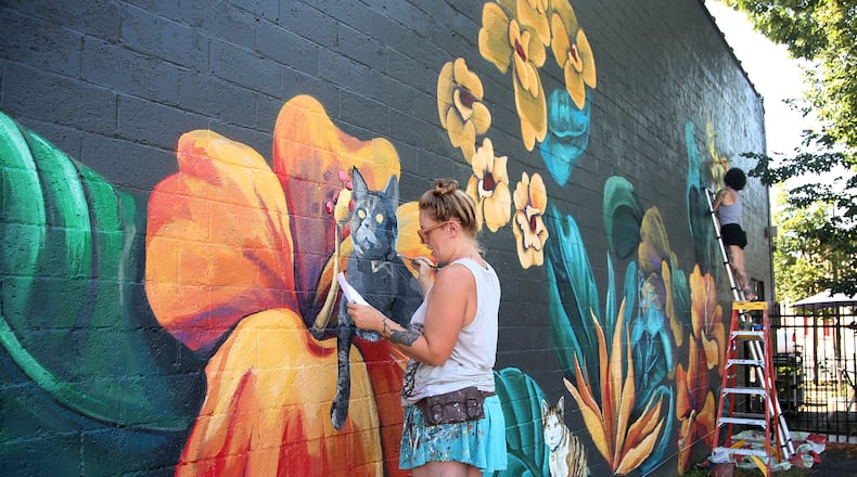 Tiffany Clark (left) and Leslea Hipp (right) of the Mural Machine is creating a mural of cats on the side of Lilyâs Bistro in Daytonâs Oregon District. People who pay a fee will be able to submit pictures of their cats for a realistic paintings on the west side of the restaurant. The project is a fundraiser for local groups dedicated to animal welfare. So far 40 cats are committed to the project and there is room for 85 feline portraits.  LISA POWELL / STAFF