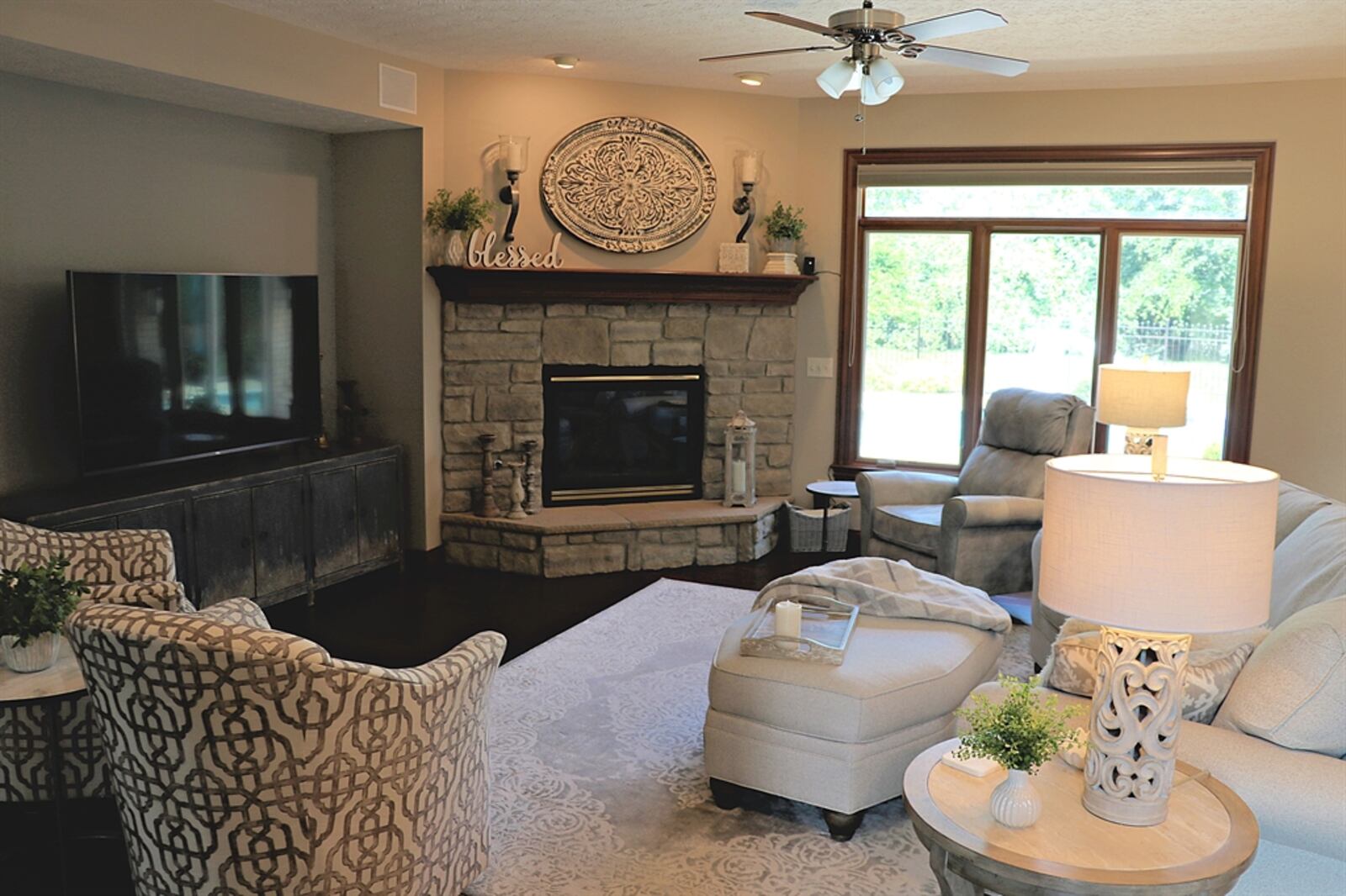 A stone gas fireplace is tucked within a corner of the great room. The fireplace has a raised stone hearth and wood-beam mantel. A furniture nook has built-in speakers above. Triple windows with a transom provide a view of the backyard pool deck.
