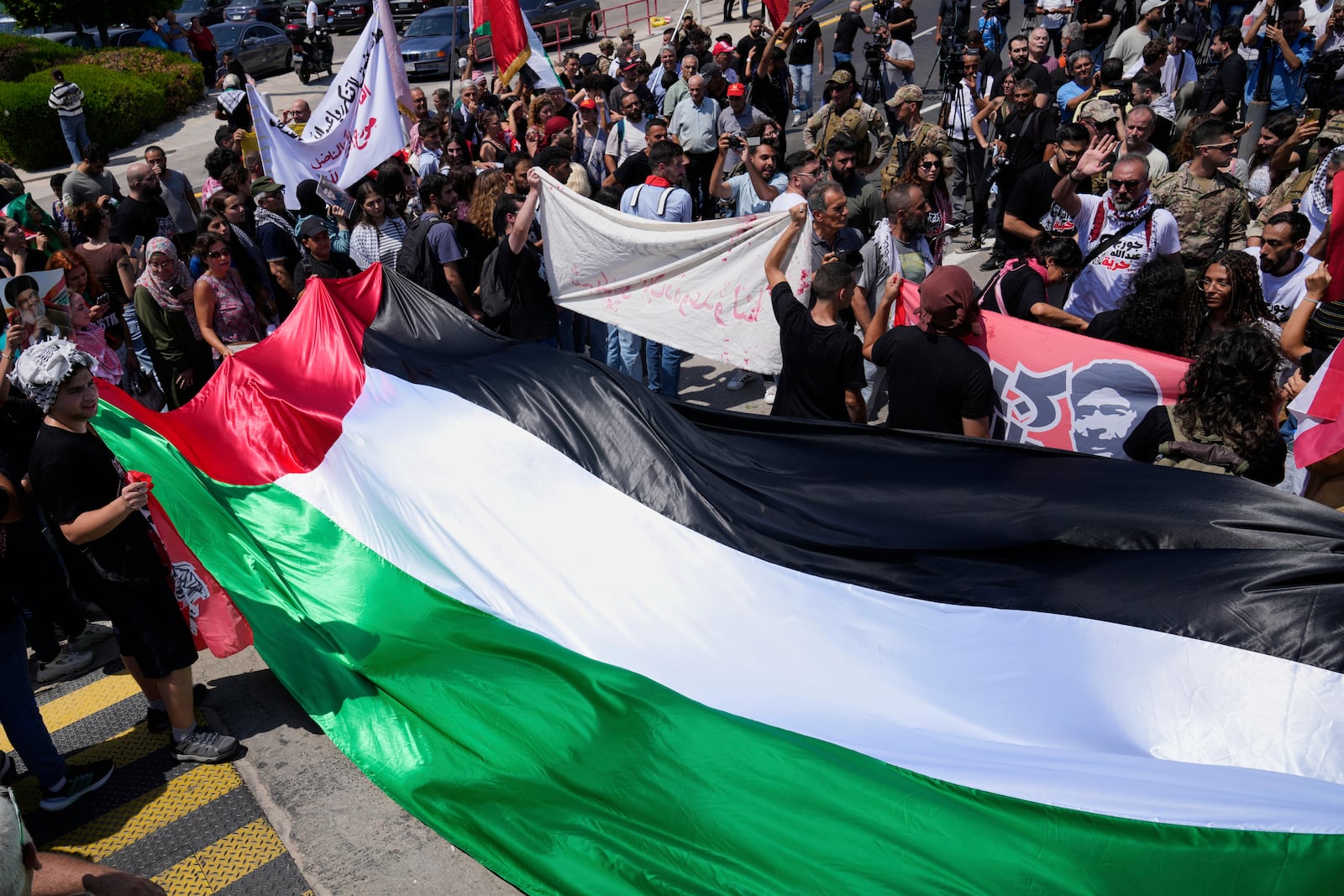 Supporters of Lebanese pro-Palestinian militant George Abdallah, who was serving a life sentence for complicity in the 1982 murders of an American and an Israeli diplomat in Paris, hold a large Palestinian flag and banners as they gather outside Rafic Hariri International Airport to welcome his return, in Beirut, Lebanon, Friday, July 25, 2025. (AP Photo/Hussein Malla)