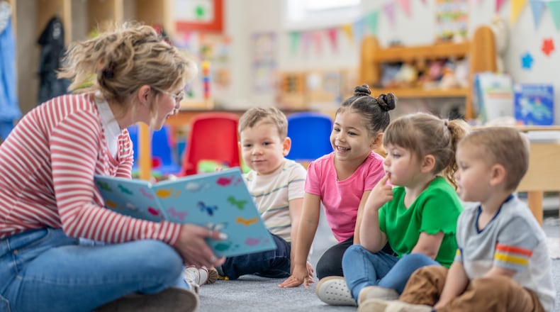 A preschool teacher sits on the floor of her classroom with a small group of students as she reads them a book. FATCAMERA/ISTOCK