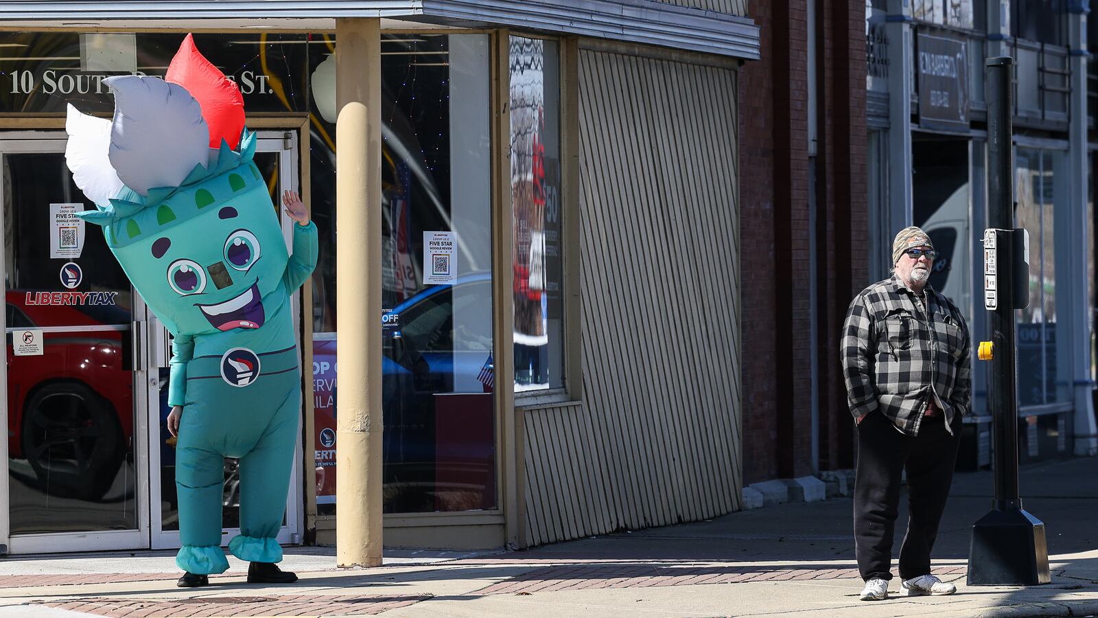 A person dressed in a mascot costume waves to traffic while standing in front of Liberty Tax in downtown Xenia  on March 24, 2025 while a man waits at a crosswalk. BRYANT BILLING / STAFF