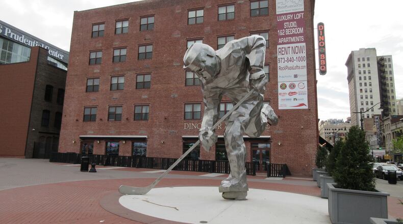 This May 11, 2017 shows "The Iron Man" sculpture of a hockey player next to the Prudential Center where the New Jersey Devils hockey team plays in Newark. In the background, signs can be seen for Dinosaur Bar-B-Cue BBQ and for Rock Plaza Lofts luxury apartments. Riots scarred Newark 50 years ago this summer, but tourism officials are hoping to attract more visitors as the city charts its comeback. (AP Photo/Beth J. Harpaz)