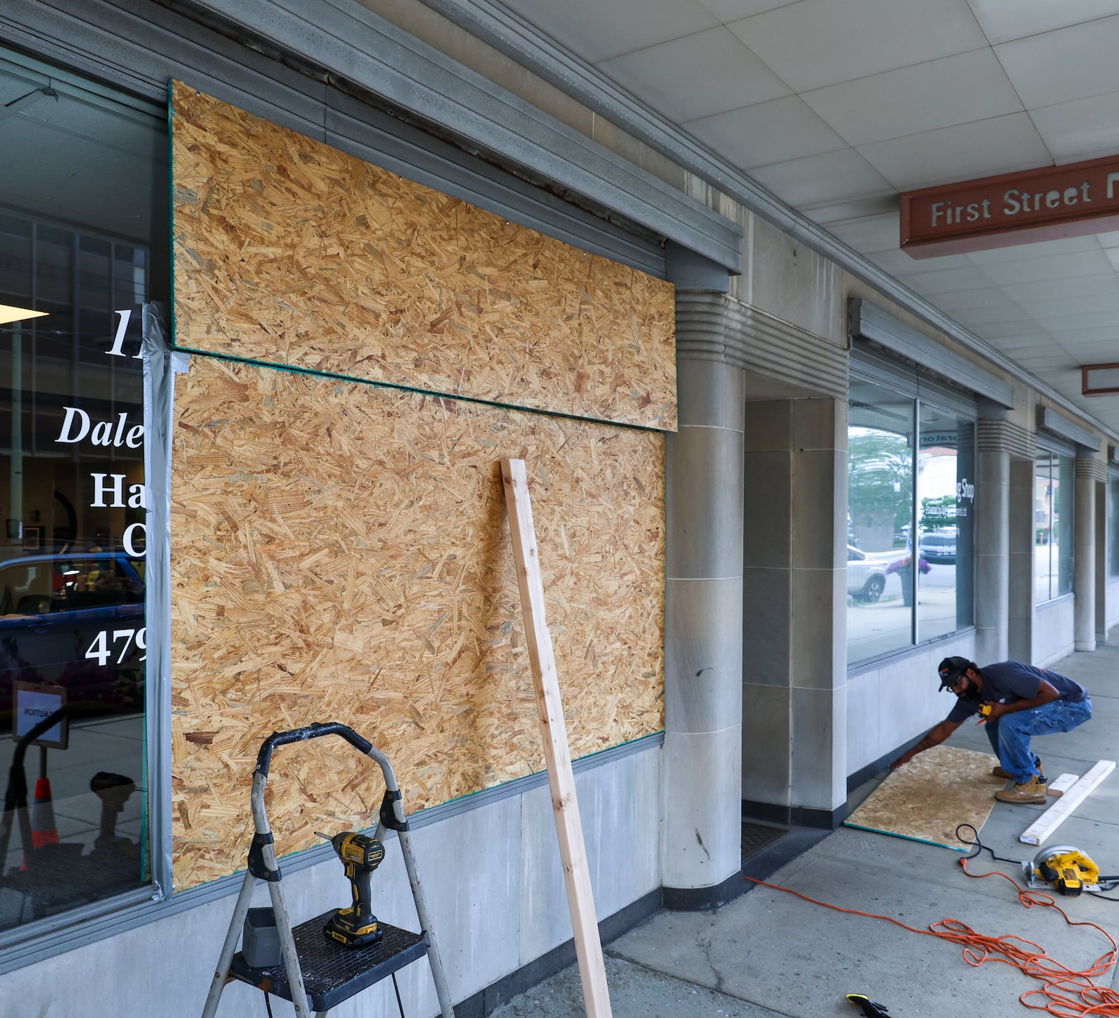 A worker cuts a wooden board to fit in a window space in front of 112 W. 1st St. in downtown Dayton. The window was damaged due to vandalism. BRYANT BILLING / STAFF