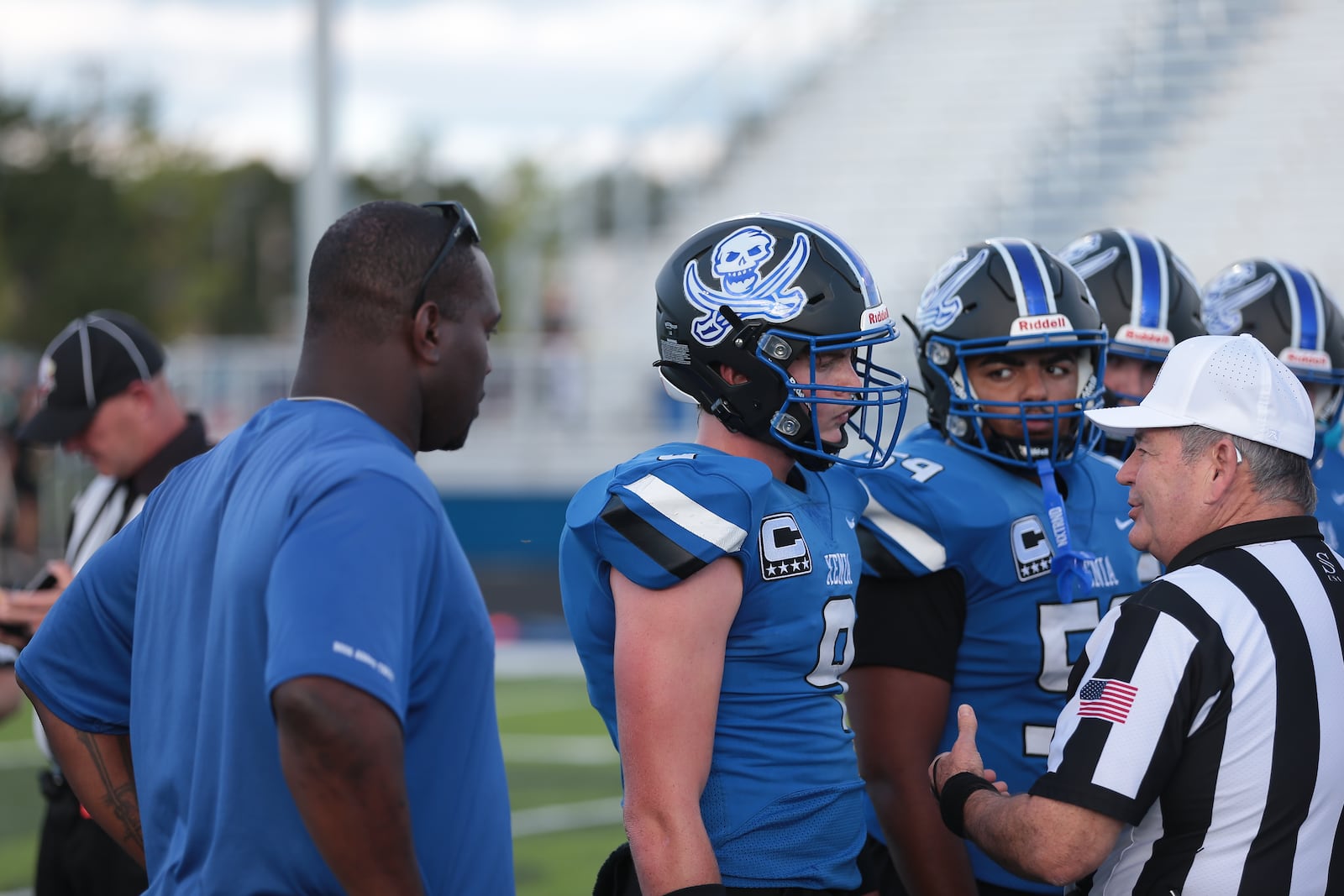 Former Xenia, University of Cincinnati and Philadelphia Eagles standout Trent Cole looks on along with Buccaneer captains Louis Freeman (9) and Kadyn Robinson (59) during the pregame coin flip Friday, Aug. 22, 2025, at Doug Adams Stadium in Xenia. Steve Black / Staff