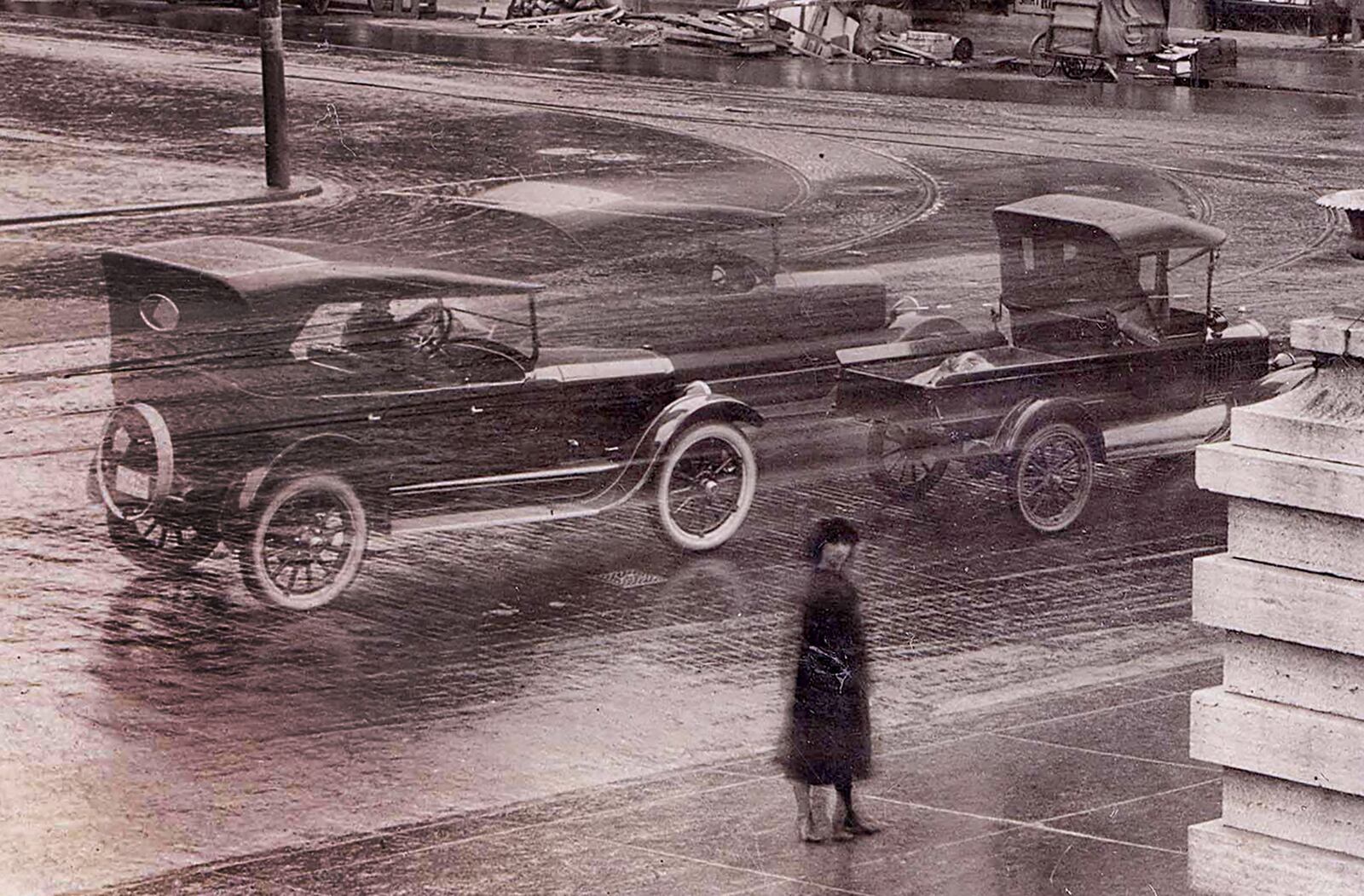 A woman standing on the sidewalk along Main Street in front of the Old Courthouse gazes at the photographer. Behind her a loaded truck and two automobiles blur by as they speed through the intersection. DAYTON METRO LIBRARY / LUTZENBERGER PICTURE COLLECTION