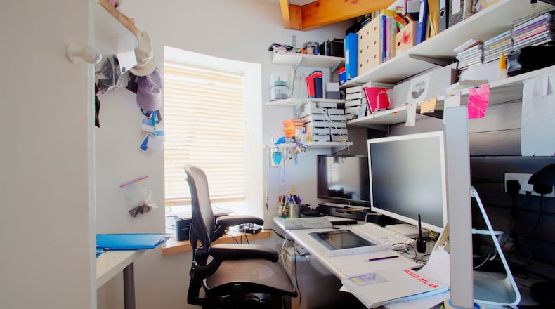 A shot of a messy desk in a home office, the room is small and cluttered, on the desk is three computer monitors and office supplies.