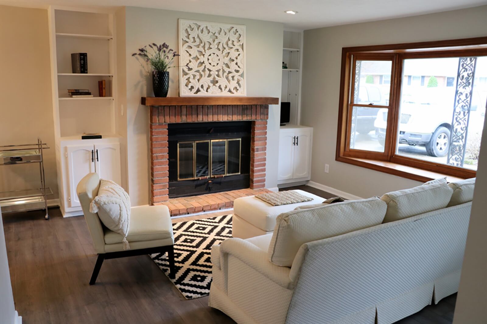 A bay window trimmed with wood fills the living room with natural light, and bricks accent the fireplace, which has a wood-beam mantel. Built-in shelves with a cabinet below flank the fireplace offering media and storage options. CONTRIBUTED PHOTO BY KATHY TYLER