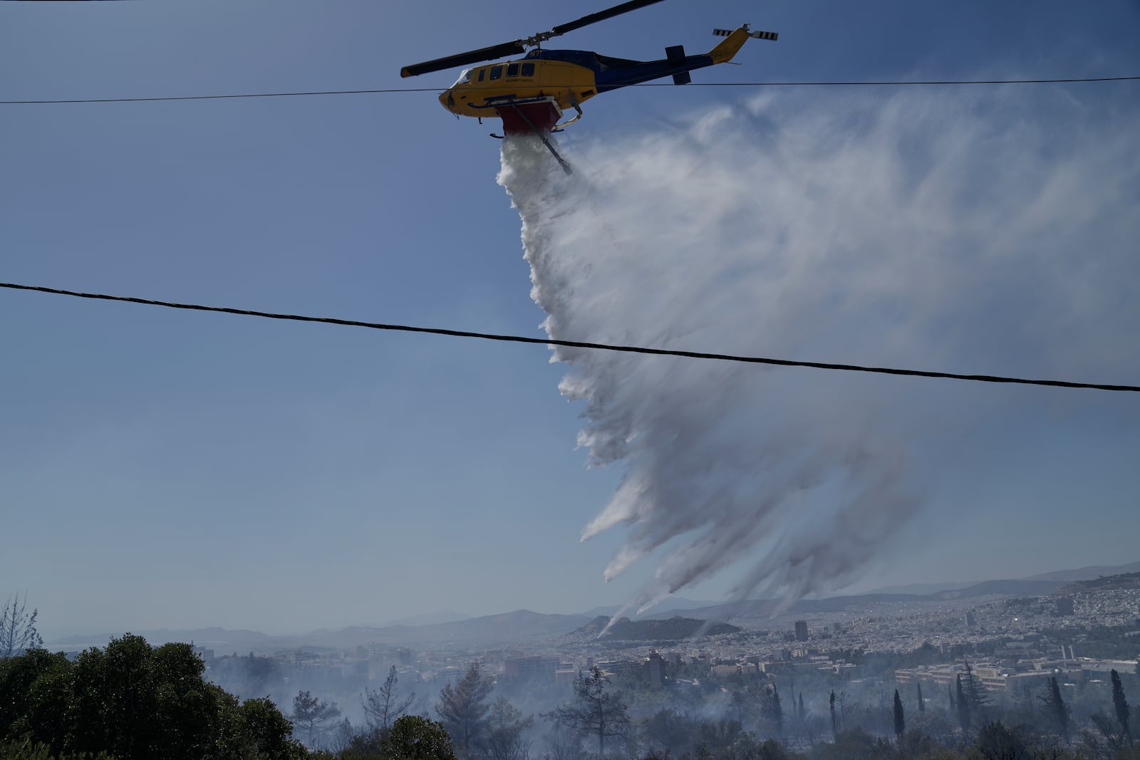 A firefighting helicopter drops water to extinguish a fire at the Polytechnic University of Athens as the Greek capital is seen in the background on Monday, July 28, 2025. (AP Photo/Thanassis Stavrakis)