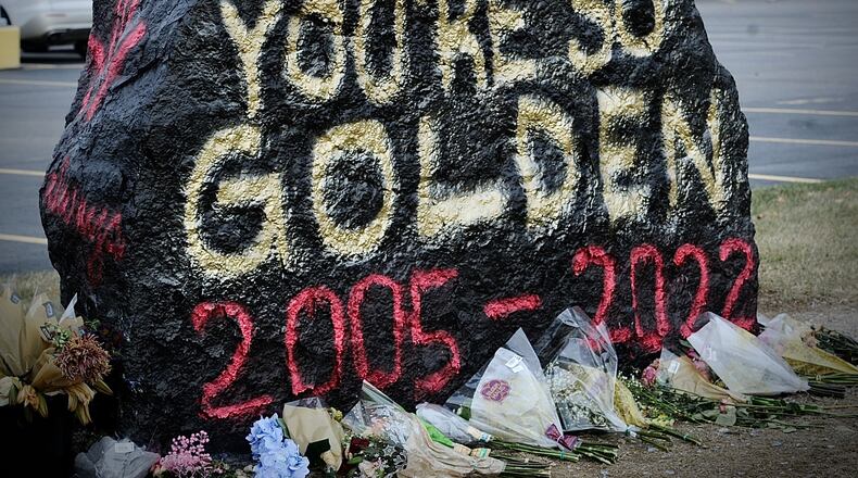 A large rock painted with "Lily, you're so golden, 2005-2022" in red and yellow spraypaint, with a memorial of flowers surrounding the base.