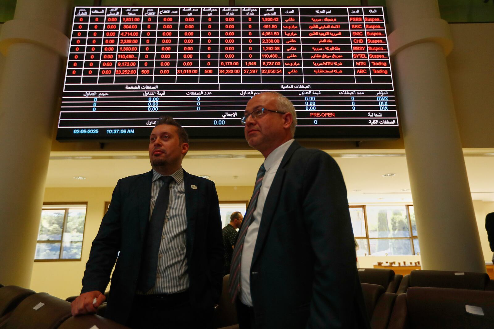 Two men stand under an electronic board showing stock prices at the Damascus Securities Exchange in Damascus, Syria, Monday, June 2, 2025. (AP Photo/Omar Sanadiki)