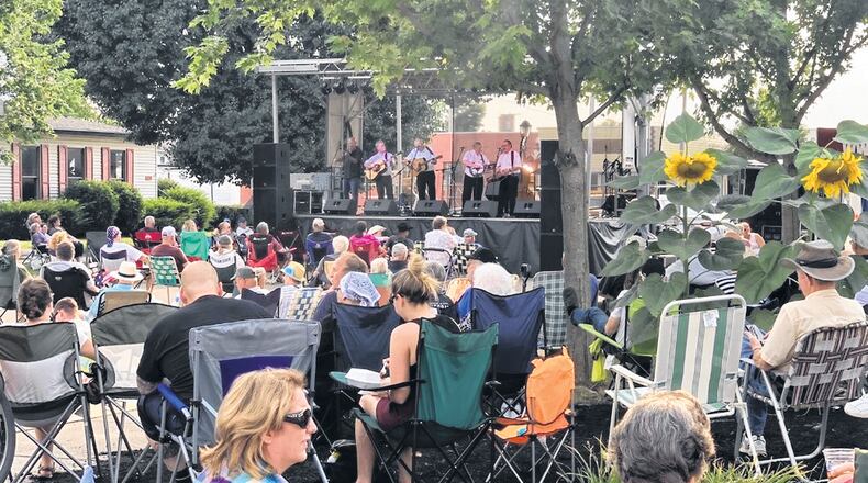 A large crowd sits outside in downtown listening to a bluegrass band on stage. Some are underneath trees, and many are sitting on lawn chairs.
