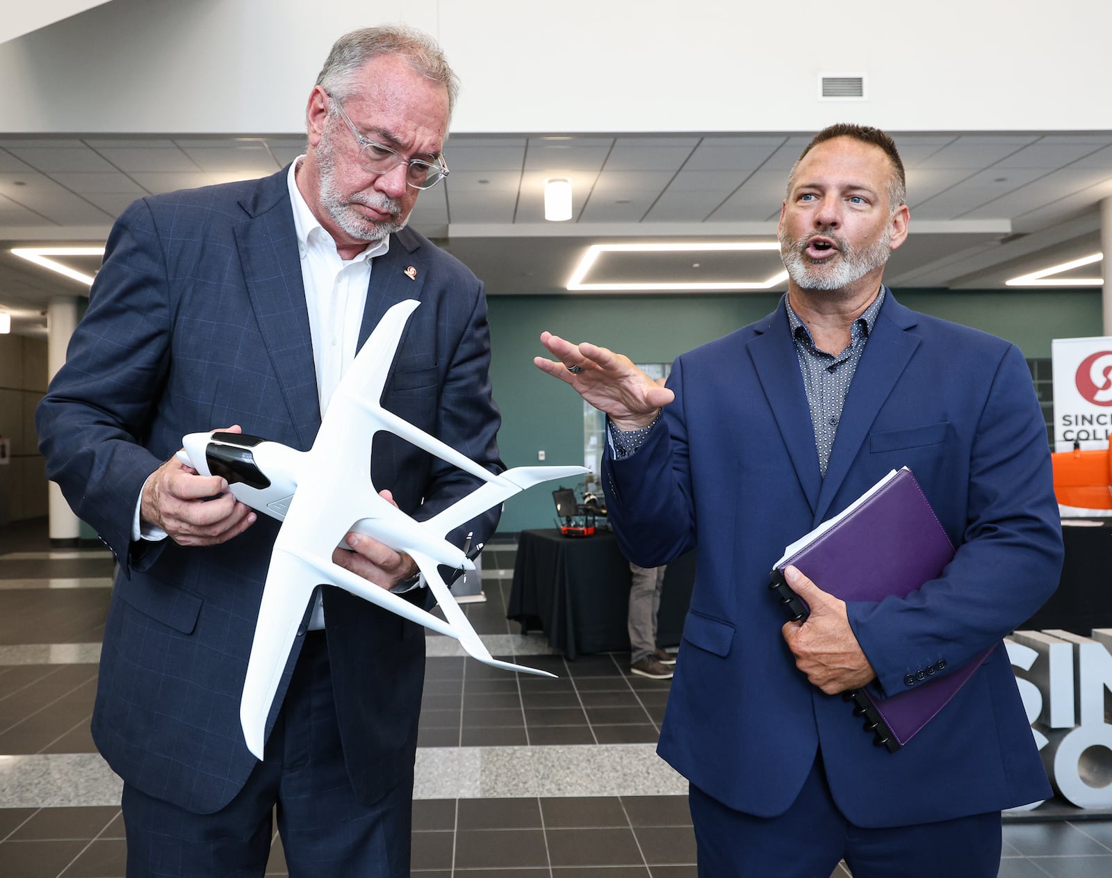 Sinclair Community College Senior Vice President Jeffery Miller talks on Wednesday, Aug. 13 at Ned J. Sifferlen Health Sciences Center as President Steven Johnson holds the model of an aircraft the college is purchasing. The aircraft is expected to be delivered in early 2026. BRYANT BILLING / STAFF