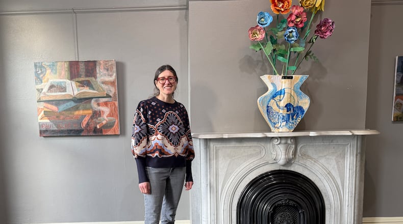 Painter Colleen Kelsey standing in her solo exhibition at Dayton Society of Artists. On the wall is "Studio Still Life" (2024-25, Oil on canvas, 28 x 30.5 inches) and on the mantle is a Delft-inspired vase and flowers made of painted cardboard, newspaper, and masking tape. HANNAH KASPER/CONTRIBUTED