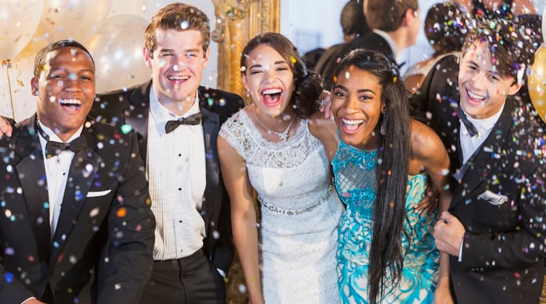 A group of five multi-ethnic teenagers and young adults dressed in formalwear - dresses and tuxedos. They are at a special event, a party or prom, standing together in a row, laughing and smiling. Or maybe a New Year's Eve party, confetti in the air to celebrate.