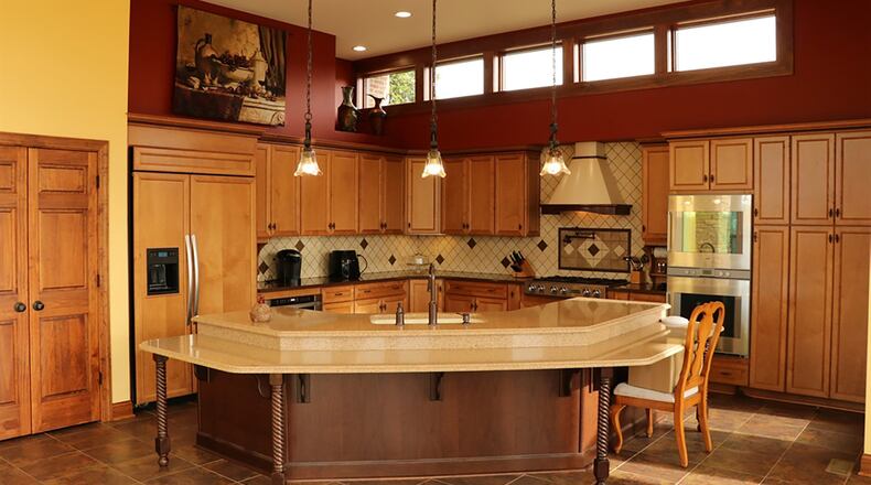 A wide, semi-circular island with a surrounding tiered seating area is placed at the entrance to the kitchen. Under a row of clerestory windows, plant shelf, recessed ceiling and hanging lights, nut-stained oversized cabinets wrap around the work space with quartz countertops and tiled backsplashes.