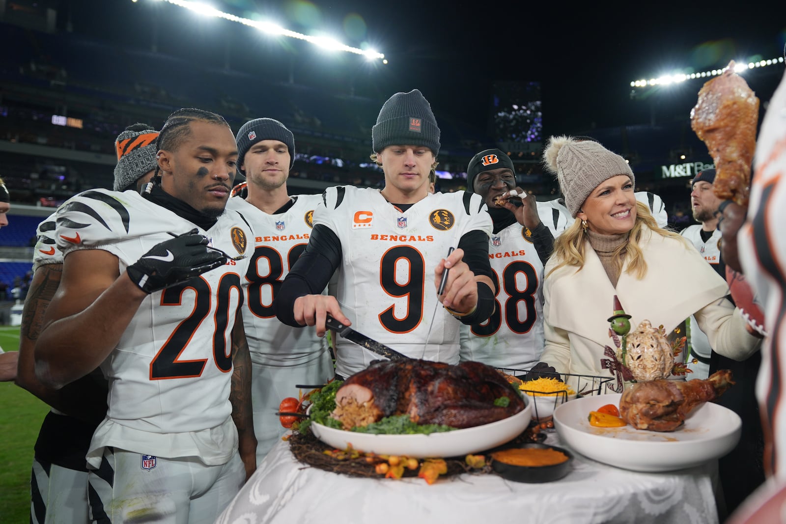 Cincinnati Bengals quarterback Joe Burrow (9) joined by NBC Sports sideline reporter Melissa Stark, right, teammates DJ Turner II (20), Mike Gesicki (88), and DJ Ivey (38) carves a turkey after NFL football game against the Baltimore Ravens, Thursday, Nov. 27, 2025, in Baltimore. (AP Photo/Stephanie Scarbrough)