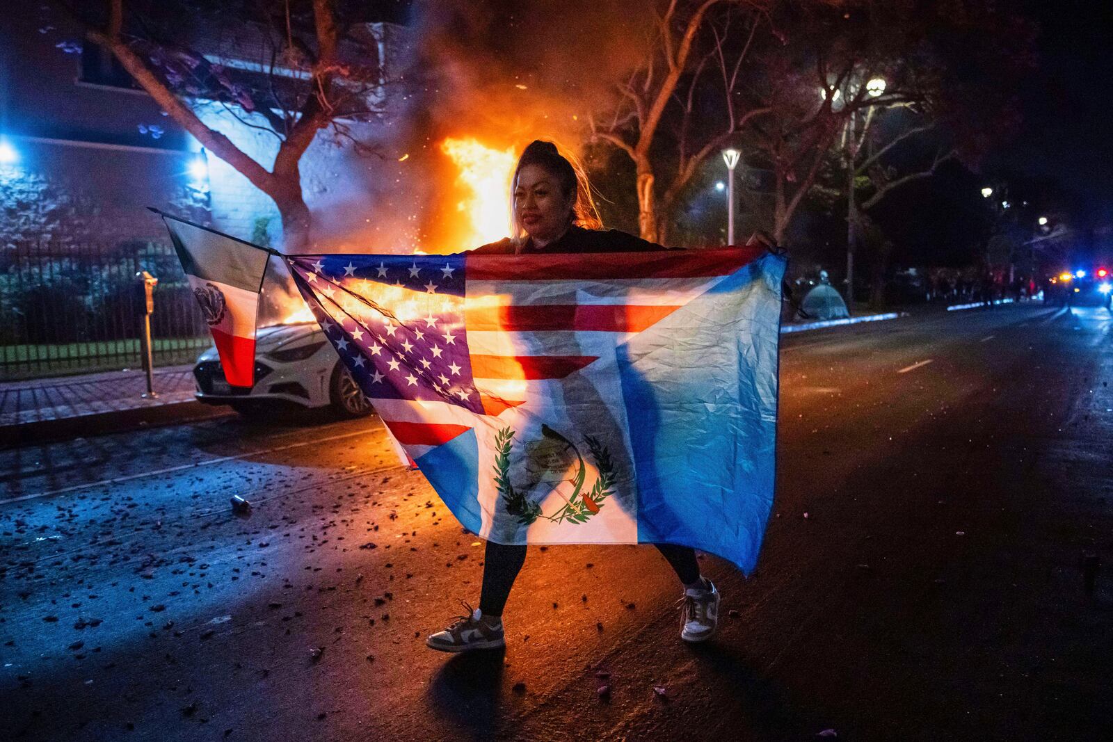 A person displaying multiple flags walks past a burning car during protests over the Trump administration's immigration raids in Los Angeles, Monday, June 9, 2025. (AP Photo/Ethan Swope)
