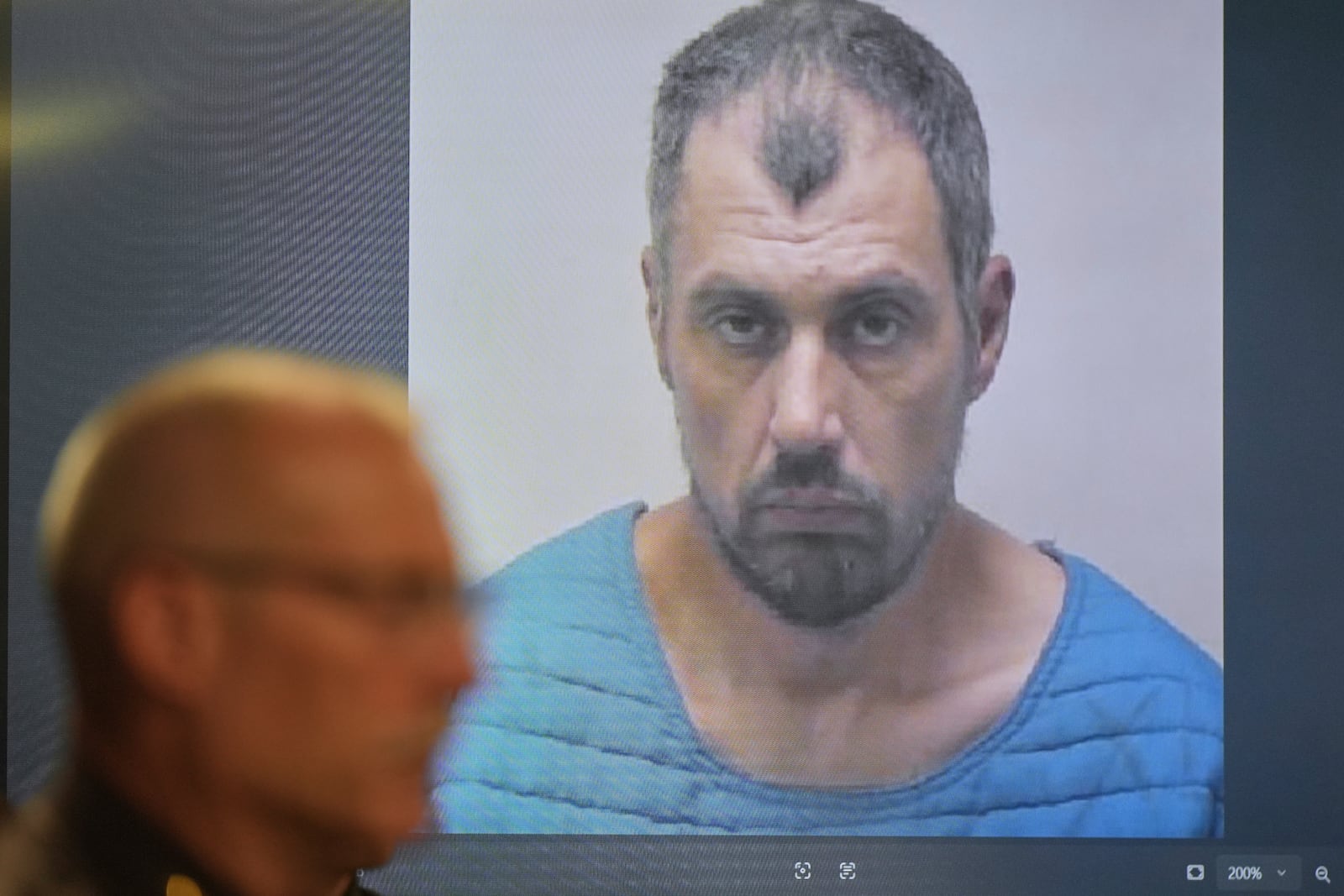 The booking photo of Bradford Gille, who is accused of stabbing 11 people at a Walmart, is seen behind Grand Traverse County Sheriff Mike Shea during a news conference Sunday, July 27, 2025, in Traverse City, Mich. (AP Photo/Ryan Sun)