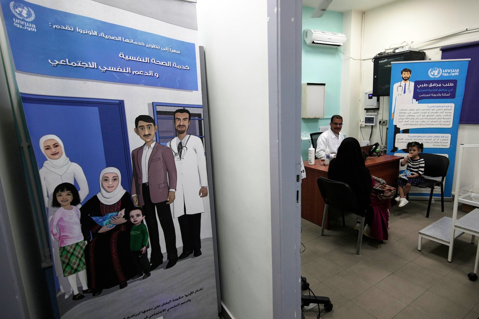 A doctor consults with a woman and her child at a medical center run by UNRWA in the Mar Elias refugee camp in Beirut, Lebanon, Monday, June 2, 2025. Posters in Arabic promote UNRWA's mental health and medical services for Palestinian refugees. (AP Photo/Hassan Ammar)