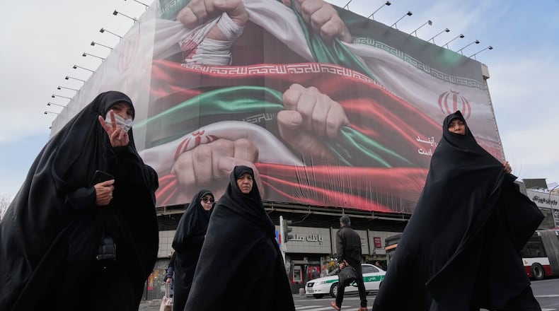 FILE - Women, one flashing a victory hand gesture, cross a street under a huge banner showing hands firmly holding Iranian national flags as a sign of patriotism, in Tehran, Iran, Jan. 14, 2026. (AP Photo/Vahid Salemi, File)
