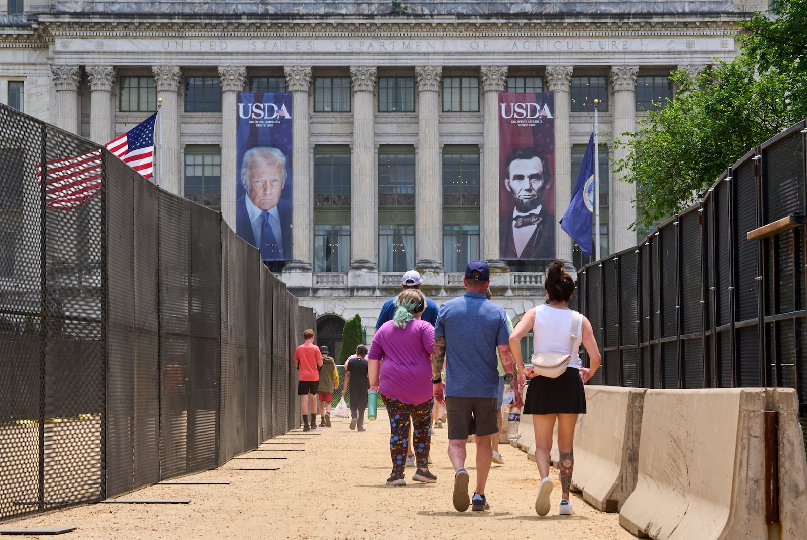 People walk in between security fencing and concrete barricades toward a large photograph of President Donald Trump and President Abraham Lincoln, draped on the USDA building, Friday, June 13, 2025, on the National Mall in Washington. (AP Photo/Jacquelyn Martin)