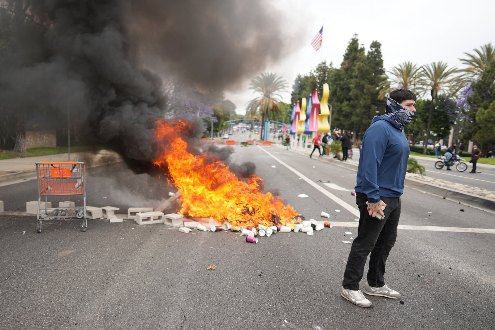 A fire burns as a protester stands across the way from Border Patrol personnel in riot gear and gas masks outside an industrial park in the city of Paramount, Calif., on Saturday, June 7, 2025 (AP Photo/Eric Thayer)