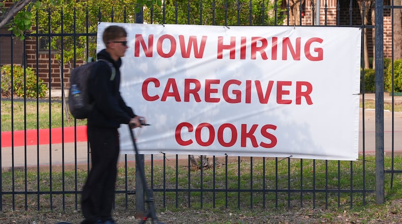 A now hiring sign sits by the sidewalk as a rider on a scooter passes in Garland, Texas, Monday, March 23, 2026. (AP Photo/LM Otero)