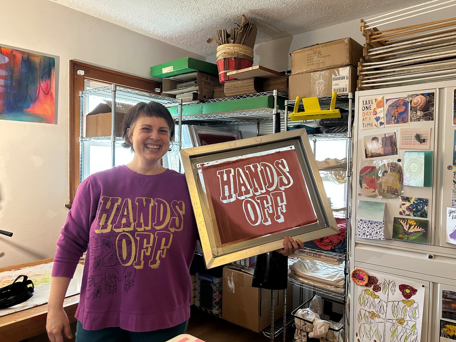 Artist Rachel Dominguez-Benner in her studio with a sample screen. In the background are cards from loved ones and her paw paw tree design.