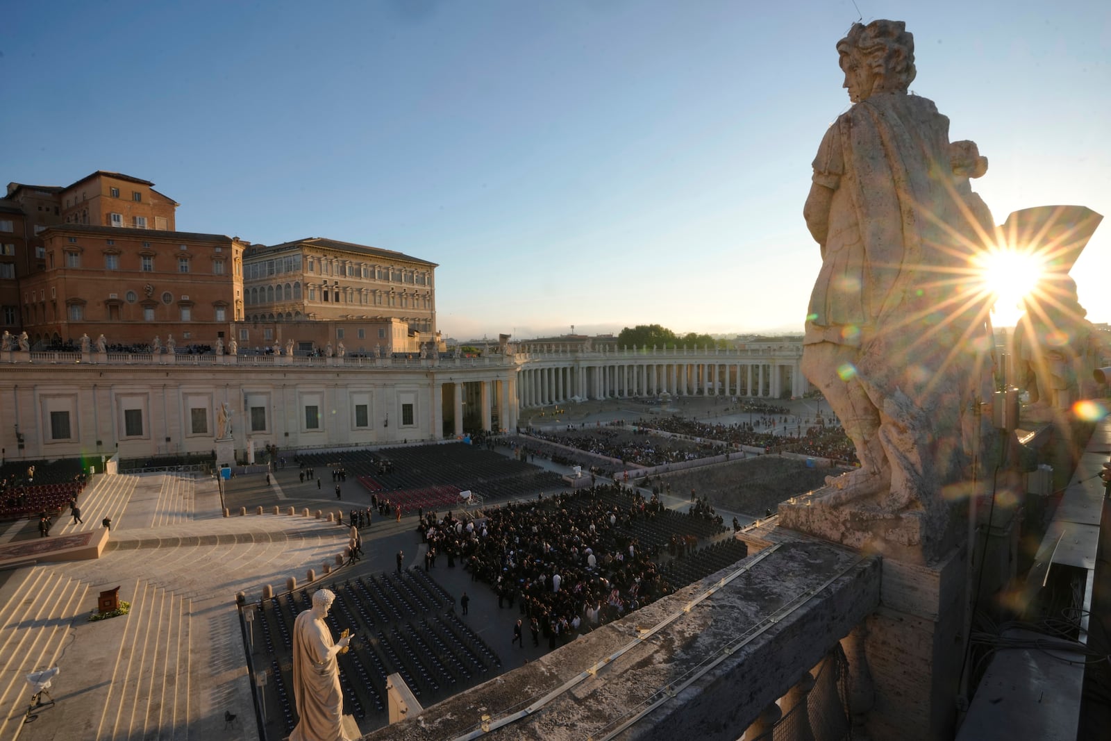 The sun rises through a statue as people begin to take their seats in St. Peter's Square, ahead of the funeral of Pope Francis at the Vatican, Saturday, April 26, 2025. (AP Photo/Markus Schreiber)