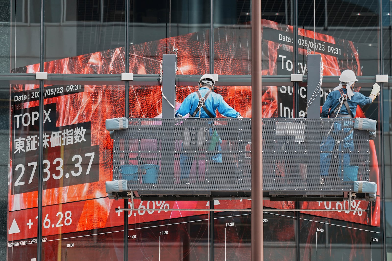 Workers clean the glass window of a building in front of an electronic stock board showing Japan's TOPIX index at a securities firm Friday, May 23, 2025, in Tokyo. (AP Photo/Eugene Hoshiko)